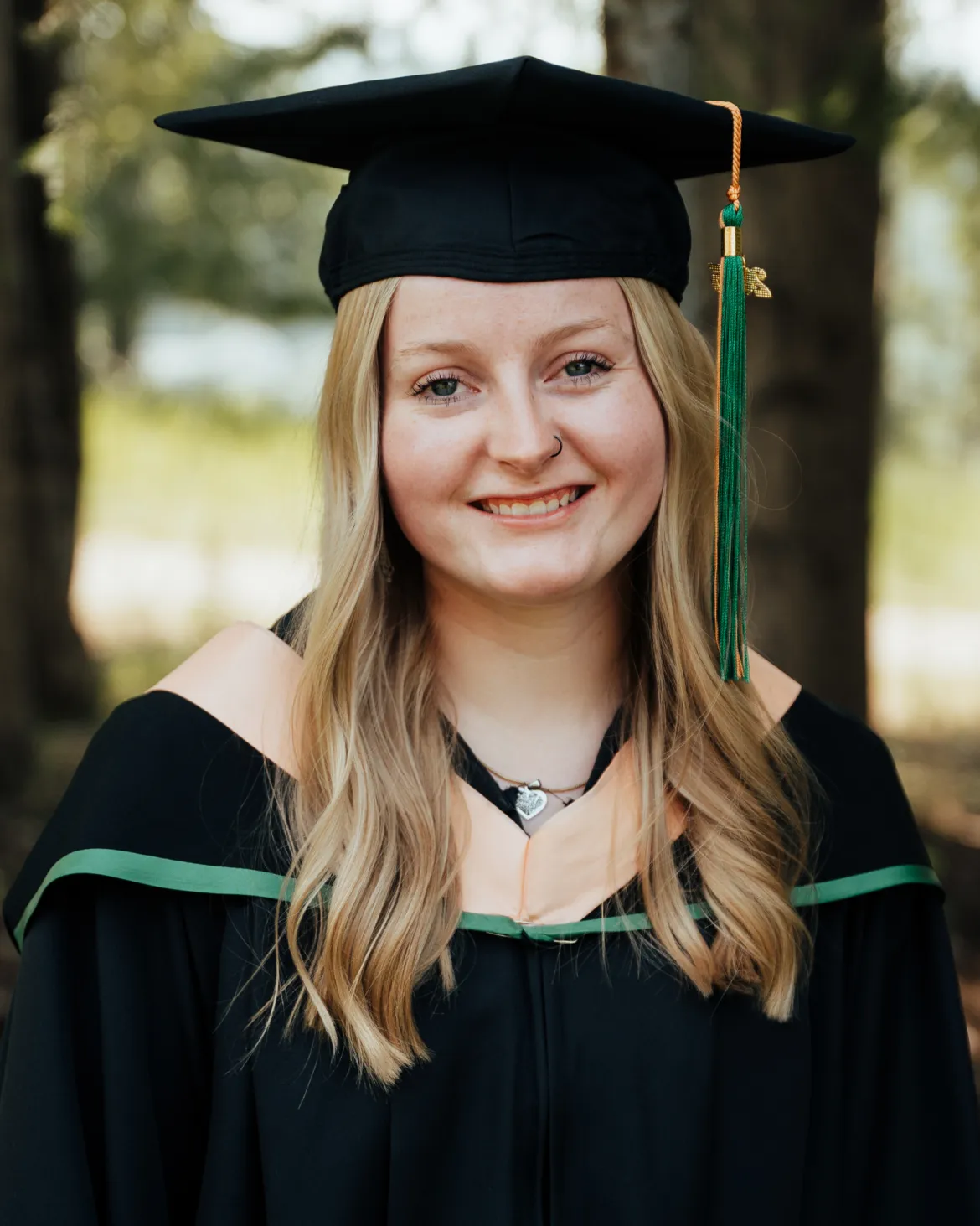 Cassidy Nikirk smiles while wearing a graduation cap and gown with green and peach trim. She is standing outdoors with trees in the background.