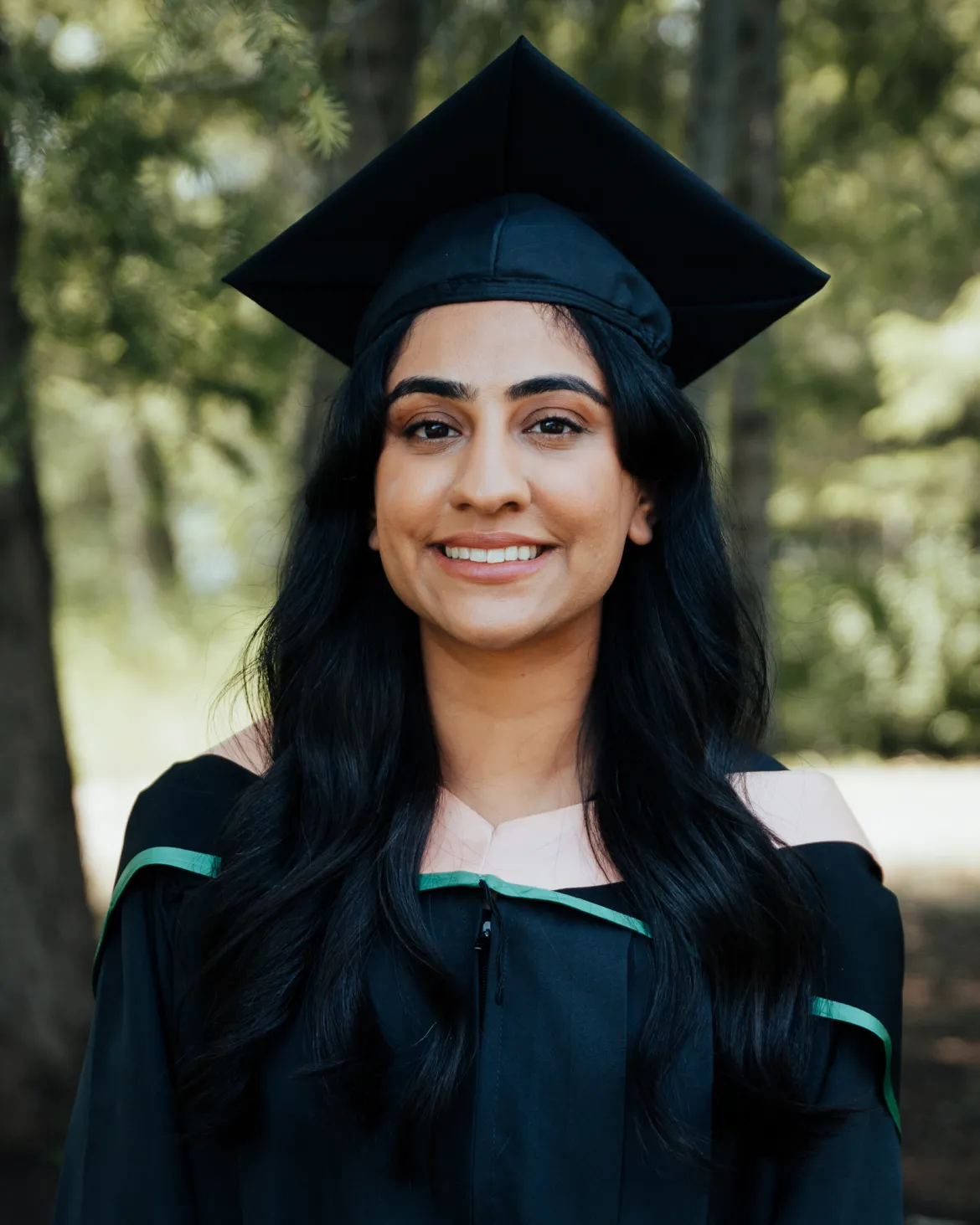Jasmine Dhesi smiles while wearing a graduation cap and gown with green and peach trim. She is standing outdoors with trees softly blurred in the background.