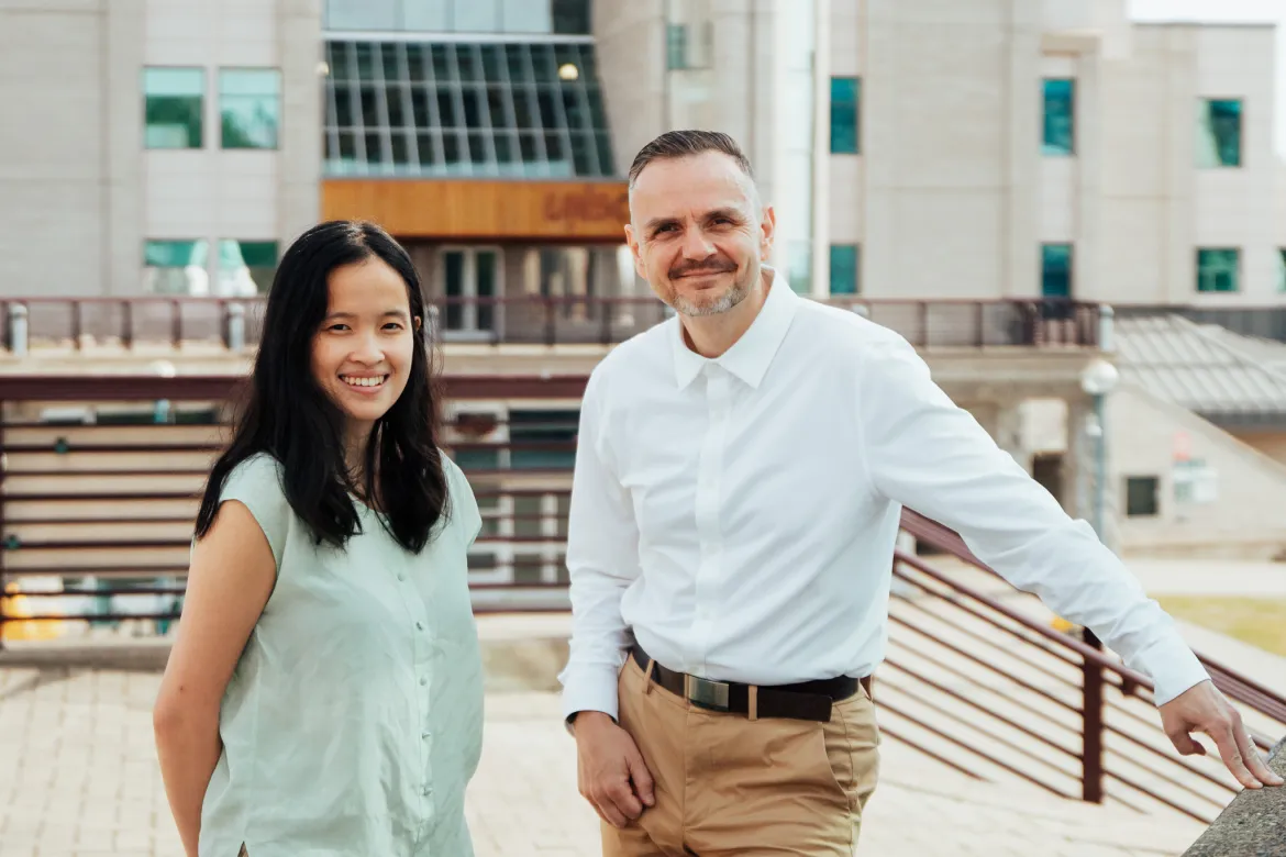 Photo shows two people standing outside at UNBC's Prince George campus. Person on right wears mint green blouse. Person on left wears white shirt and brown pants.