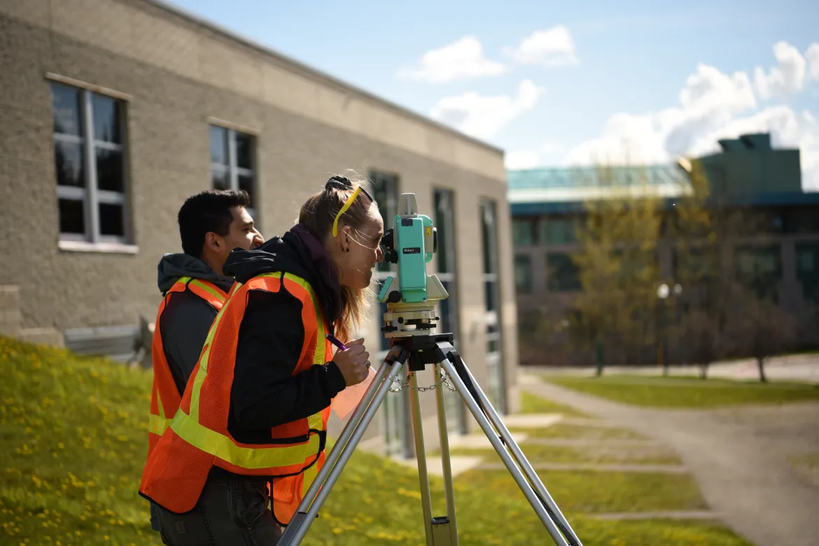 Two students in safety vests operate surveying equipment.