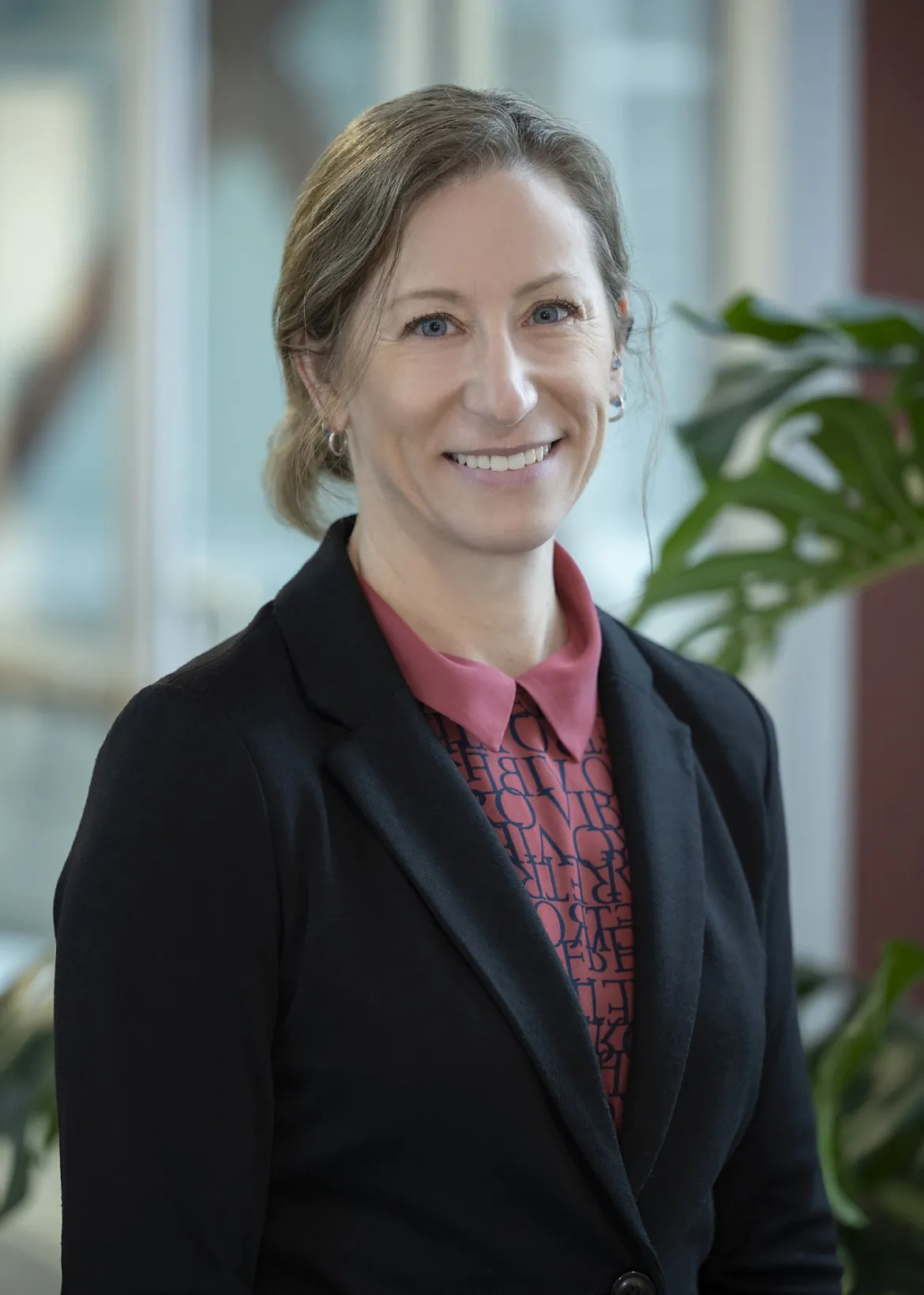 Person wearing rose-coloured blouse with pattern and dark blazer poses for photo with greenery in background.