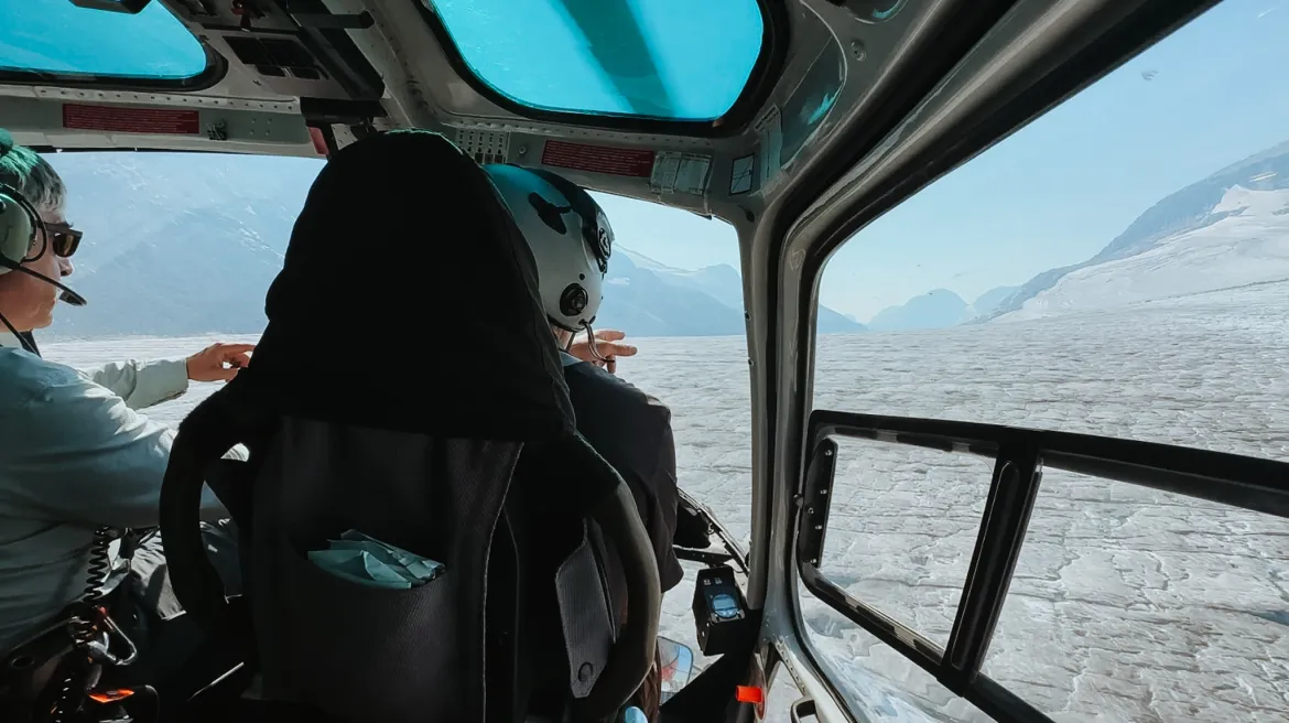 View from behind of two people inside a helicopter looking out and pointing at a glacier.