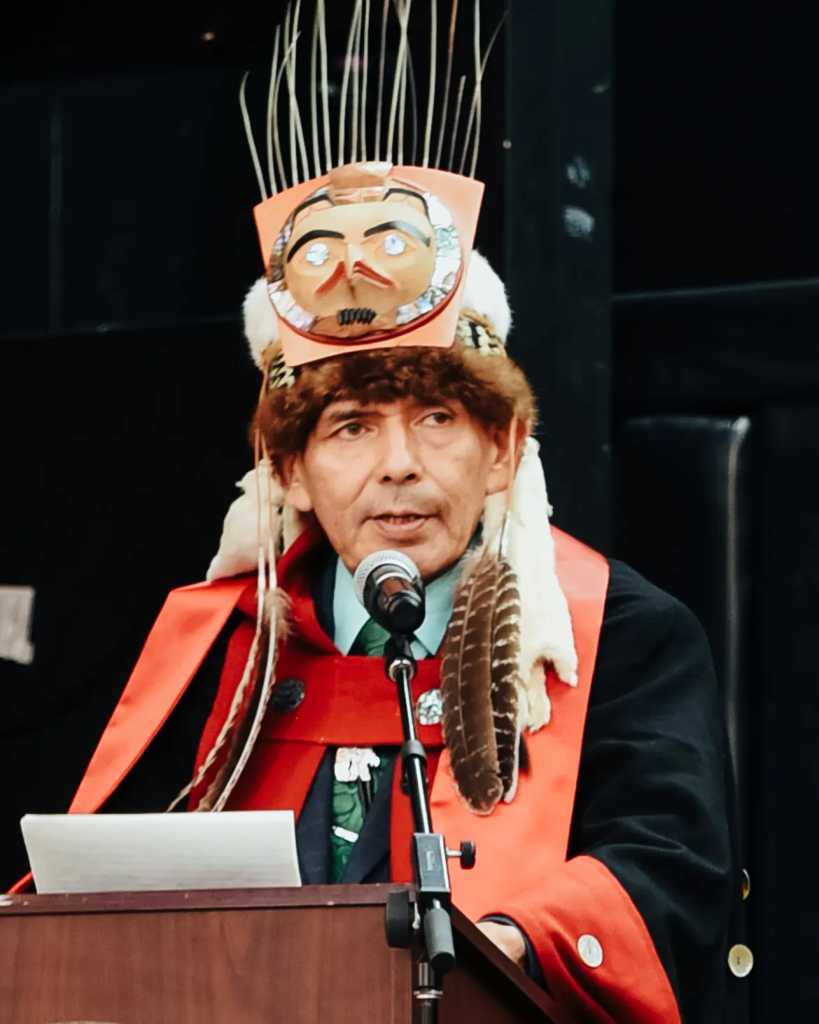 Person wearing red and black traditional regalia stands at a podium.
