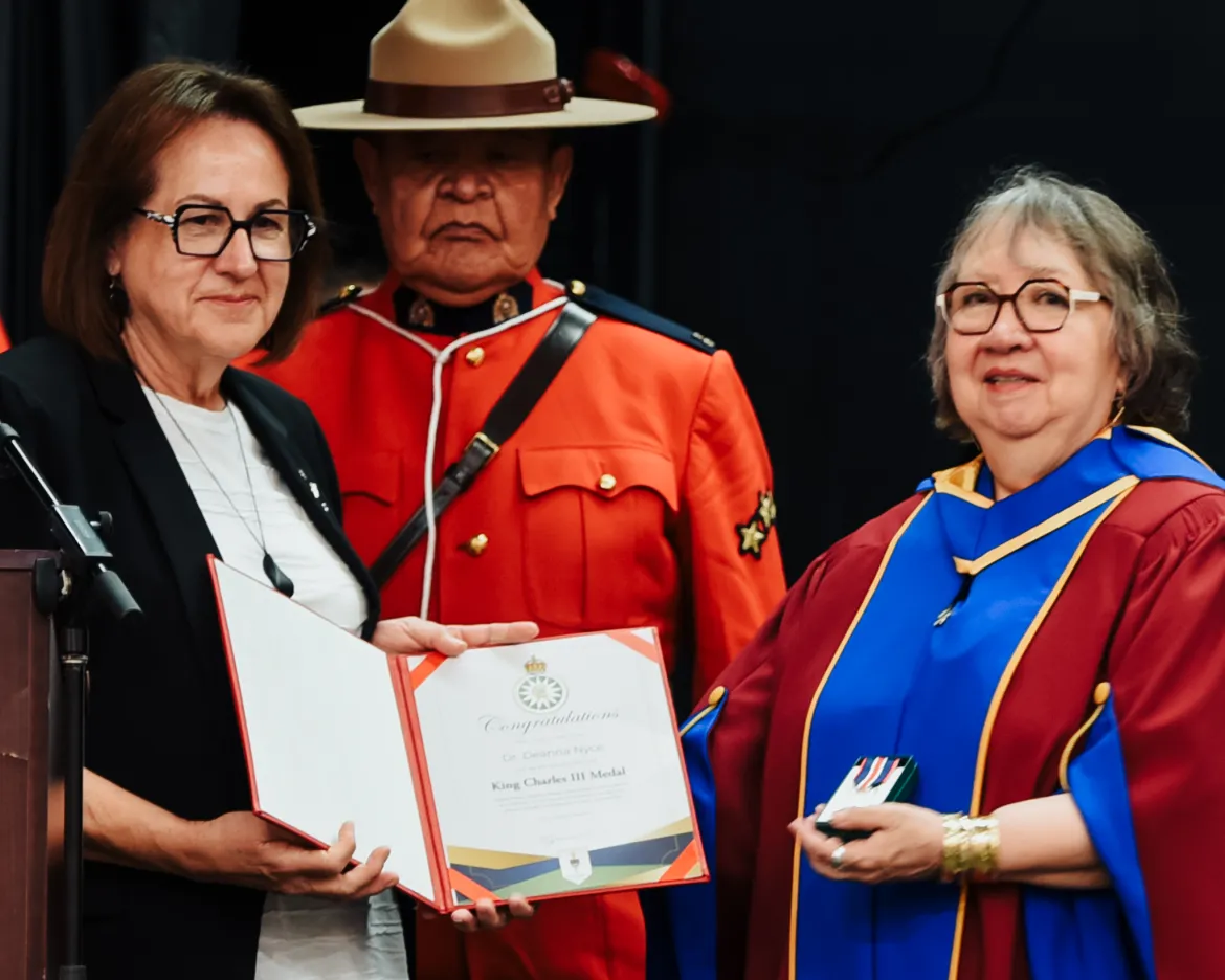 Two people stand in foreground. Person on left wears white blouse and black blazer. Person on right wears red and blue academic regalia. RCMP officer in red serge in background.