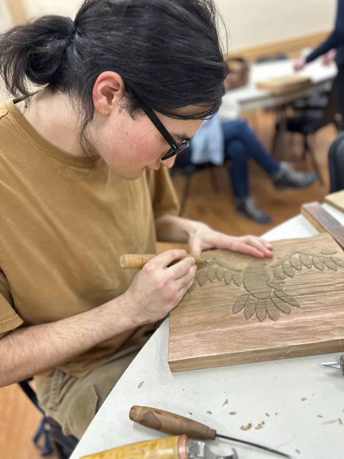 Brent Elias Man in tan t-shirt carves wooden plank at a table