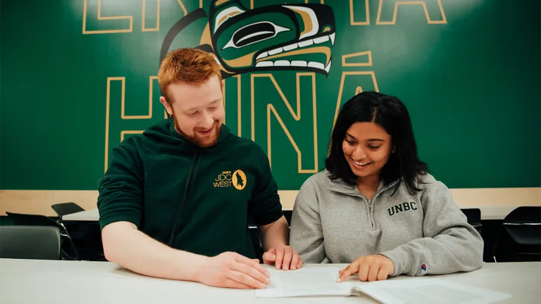 Two students with UNBC Logo in the back, male student with red hair and female student with darker skin and black hair