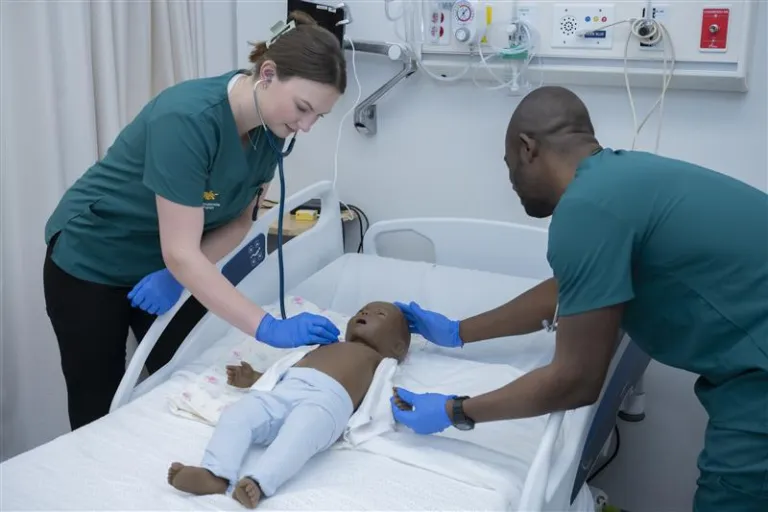 Two nursing students standing around a nursing bed, on either side of a toddler-sized mannequin, checking vitals. 