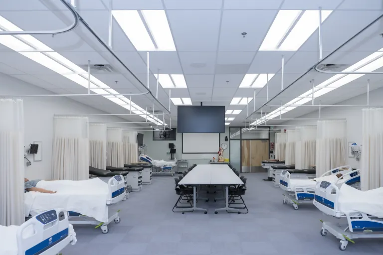 Full view of simulation lab, patient beds divided by curtains and a conference table in the middle of the room, facing a projector screen. 