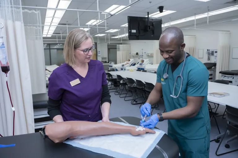 Student practicing administering intravenous medicine under guidance of Instructor, on a simulation arm. 