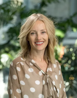 Person wearing beige blouse with white polka dots with greenery in background.