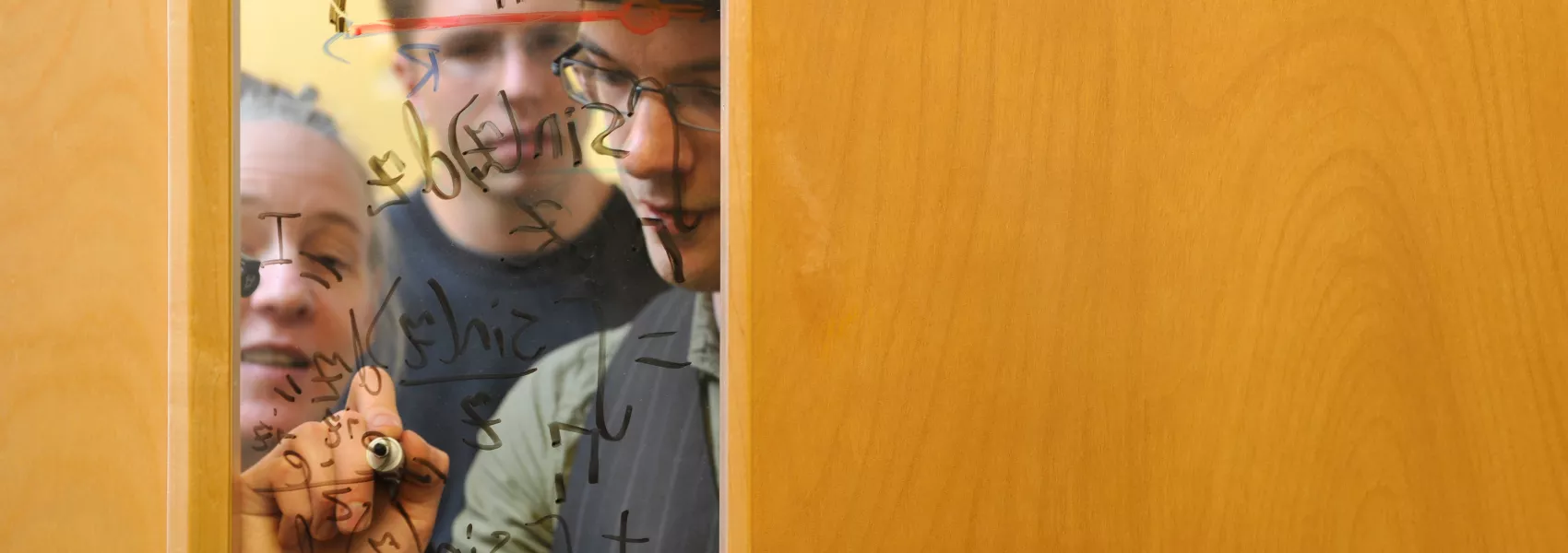 A student writes a math equation on a window of a classroom as two others look on.